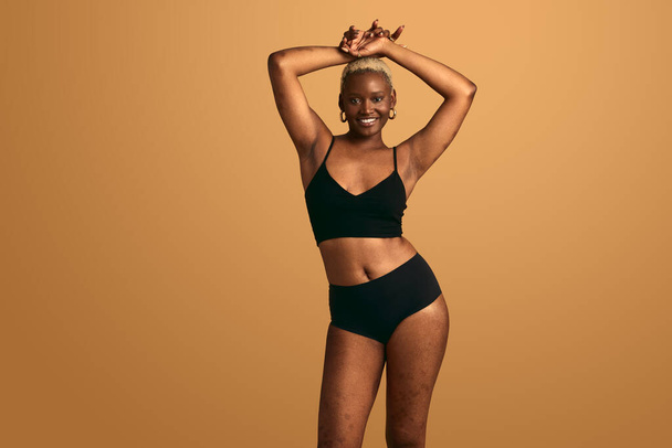 Positive young African American female model in black lingerie looking at camera and smiling while standing against beige background with raised arms in studio - Φωτογραφία, εικόνα