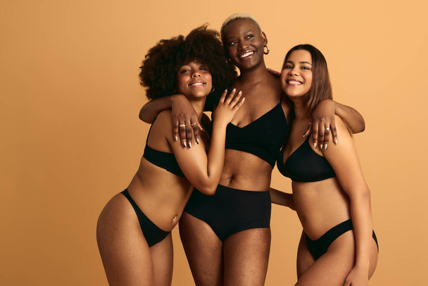 Self assured young multiracial plus size female models in black lingerie embracing and smiling against beige background - Fotografie, Obrázek