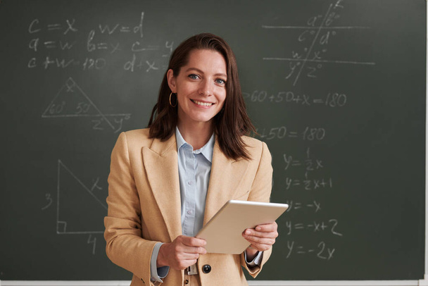 Waist up portrait of young female teacher looking at camera and smiling while standing by blackboard in school classroom, copy space - Photo, image
