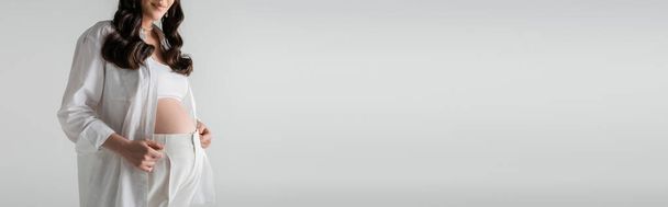 vista ritagliata di donna sorridente e incinta in camicia bianca alla moda e pantaloni in piedi isolato su sfondo grigio, concetto di moda maternità, banner - Foto, immagini