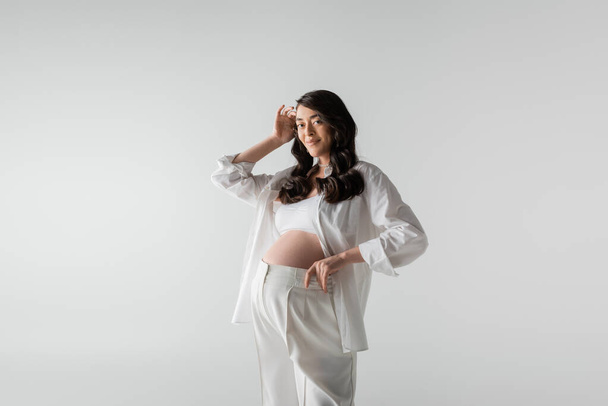 fashionable pregnant woman with wavy brunette hair posing in white pants and shirt while smiling at camera isolated on grey background, maternity style concept - Photo, Image