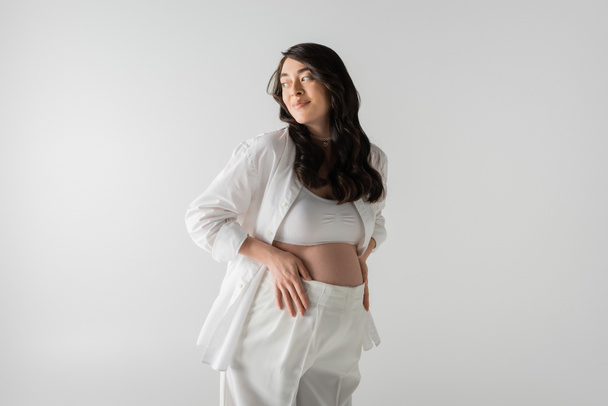 femme enceinte gaie en chemise élégante blanche, haut de culture et pantalon touchant le ventre et regardant loin isolé sur fond gris, concept de mode maternité - Photo, image