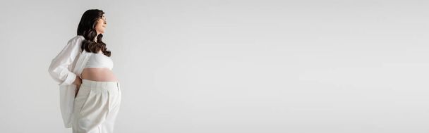 vista laterale della modella incinta con capelli castani ondulati in posa bianca camicia elegante e pantaloni isolati su sfondo grigio, concetto di moda maternità, banner, moda maternità  - Foto, immagini