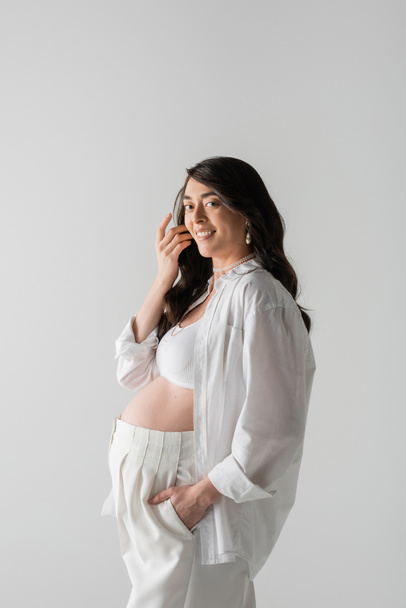 encantadora mujer embarazada con el pelo ondulado morena, en camisa blanca, top de la cosecha y pantalones, sonriendo y mirando a la cámara aislada sobre fondo gris, concepto de moda de maternidad - Foto, Imagen