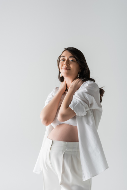 joyful mother-to-be with wavy brunette hair touching neck and looking at camera while posing in white shirt and pants isolated on grey background, maternity style concept, pregnant woman  - Фото, изображение