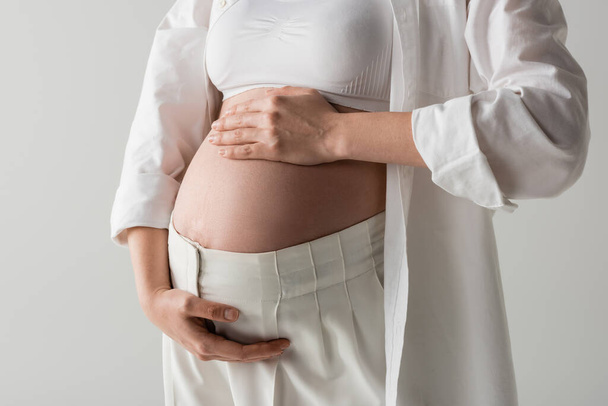 μερική άποψη της μελλοντικής μητέρας σε λευκό κομψό πουκάμισο, κορυφή καλλιέργεια και παντελόνι τρυφερά αγκαλιάζει την κοιλιά απομονώνονται σε γκρι φόντο, έννοια της μόδας μητρότητας, έγκυος γυναίκα  - Φωτογραφία, εικόνα