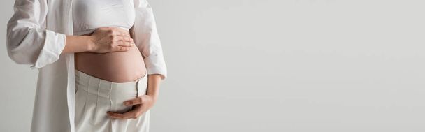 częściowy widok przyszłej matki w białej bluzce, koszuli i spodniach obejmującej brzuch, stojącej na szarym tle, koncepcja mody macierzyńskiej, baner, kobieta w ciąży  - Zdjęcie, obraz