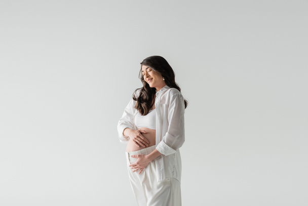 modelo embarazada atractiva y elegante en top blanco de la cosecha, camisa y pantalones abrazando barriga y sonriendo con los ojos cerrados aislados sobre fondo gris, concepto de moda de maternidad - Foto, imagen