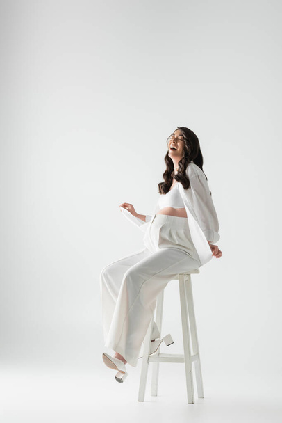 full length of excited pregnant woman with wavy brunette hair sitting on stool in white trendy clothes and smiling with closed eyes on grey background, maternity fashion concept - Photo, Image
