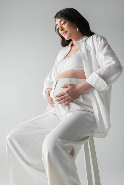smiling and appealing pregnant woman in white shirt, crop top and pants sitting on stool and embracing tummy isolated on grey background, maternity style concept, mother-to-be - Fotó, kép