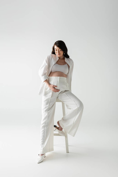 full length of happy mom-to-be in white pants, crop top and shirt posing near stool and touching tummy on grey background, fashionable pregnancy concept, pregnant woman  - Фото, зображення