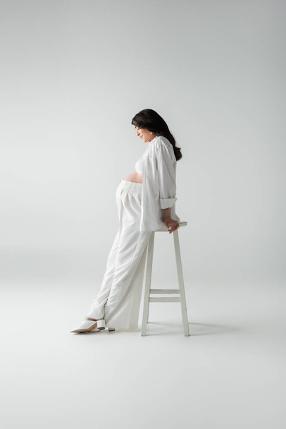 side view of mother-to-be with wavy brunette hair posing near stool in white pants, crop top and shirt near stool on grey background, pregnancy fashion concept - Фото, зображення