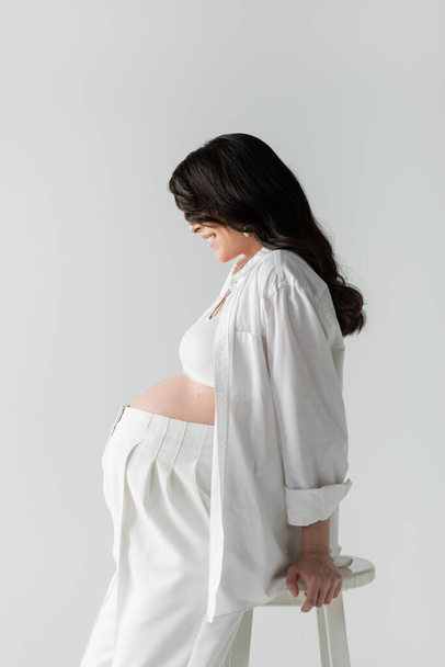 side view of happy pregnant woman with wavy brunette hair posing in white crop top, shirt and pants isolated on grey background, fashionable maternity concept  - Fotó, kép