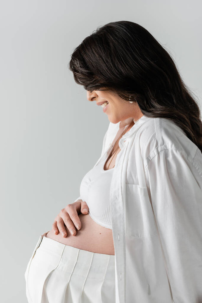 вид сбоку улыбающейся беременной женщины в белом топе и рубашке, лицо затемнено волнистыми волосами брюнетки, касание живота изолировано на сером фоне, концепция моды для беременных - Фото, изображение