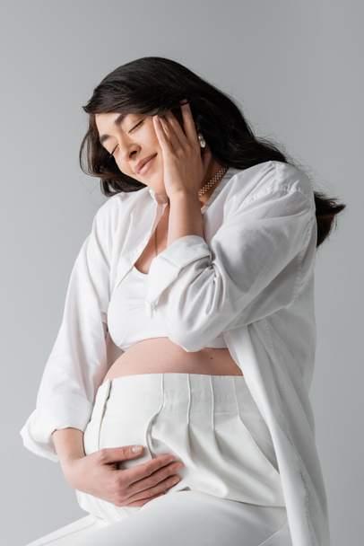brunetka těhotná žena v bílé plodině top, košile a kalhoty dotýkat břicho a šťastný obličej se zavřenýma očima izolované na šedém pozadí, mateřství koncepce - Fotografie, Obrázek