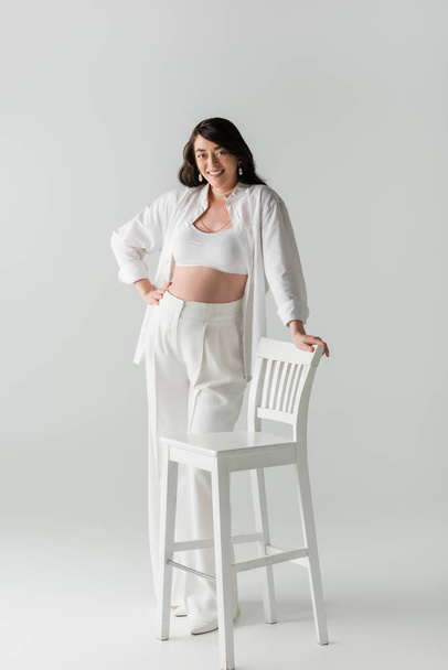 full length of charming and cheerful mom-to-be in trendy pants, crop top and shirt posing with hand on hip near white chair on grey background, maternity fashion concept, pregnant woman  - Fotó, kép