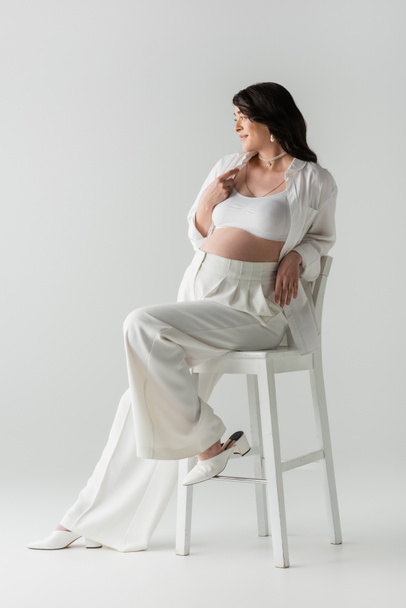 full length of trendy pregnant woman in white crop top, shirt and pants sitting on chair, smiling and looking away on grey background, maternity fashion concept - Photo, Image