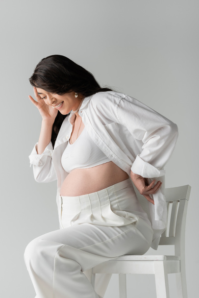 joyful mom-to-be sitting on chair in white crop top, shirt and pants while holding hand near happy face isolated on grey background, maternity fashion concept - Fotografie, Obrázek