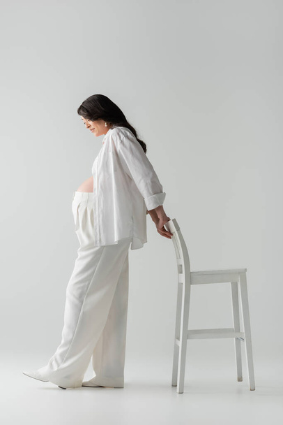 side view of brunette and stylish pregnant woman in white pants, shirt and crop top posing with chair and smiling on grey background, maternity fashion concept - Fotografie, Obrázek