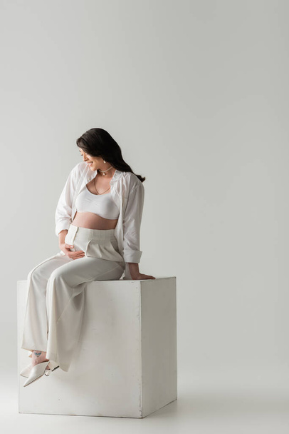 full length of smiling mother-to-be in stylish pants, crop top and shirt touching tummy while sitting on white cube on grey background in studio, maternity fashion concept, pregnant woman  - Foto, immagini
