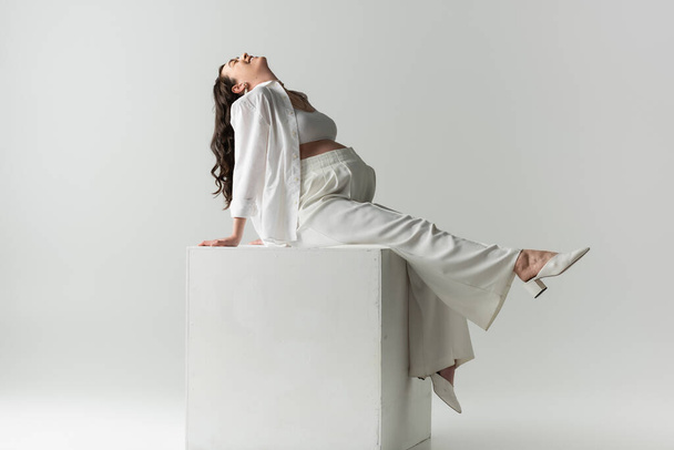 full length of excited future mother in white stylish clothes such as pants, crop top and shirt sitting on white cube on grey background, maternity fashion concept - Fotografie, Obrázek