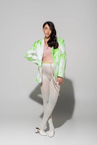 full length of pretty brunette mother-to-be in green and white jacket, crop top, beads belt and tights posing with hand on hip and looking at camera on grey background, trendy pregnancy concept - Photo, Image
