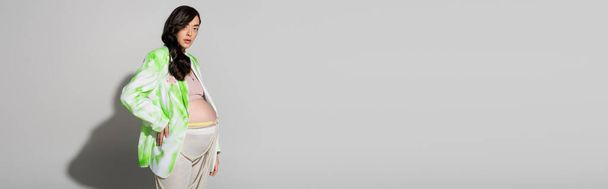 стильная будущая мама с волнистыми волосами брюнетки, в зеленой и белой куртке, леггинсы и бусы пояса стоя с рукой на бедре на сером фоне, концепция моды для беременных, баннер, беременная женщина  - Фото, изображение