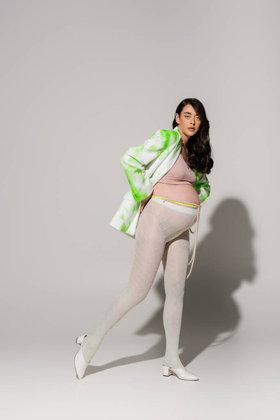 full length of brunette and charming pregnant woman in leggings, crop top, green and white jacket posing on grey background, maternity fashion concept, expectation  - Foto, Imagem