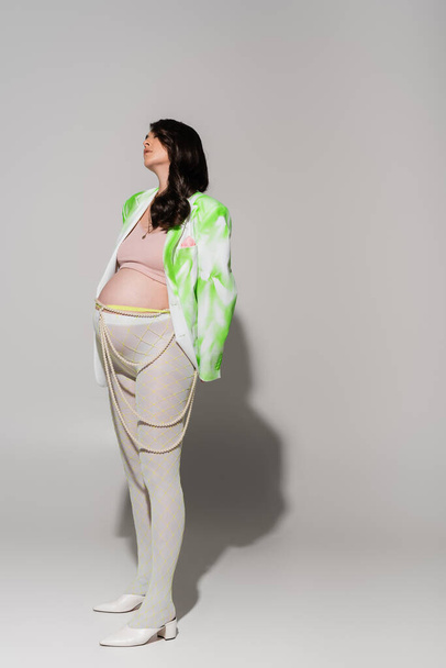 full length of fashionable pregnant woman in tights, crop top, green and white jacket and beads belt standing on grey background, maternity fashion concept, expectation  - Photo, Image