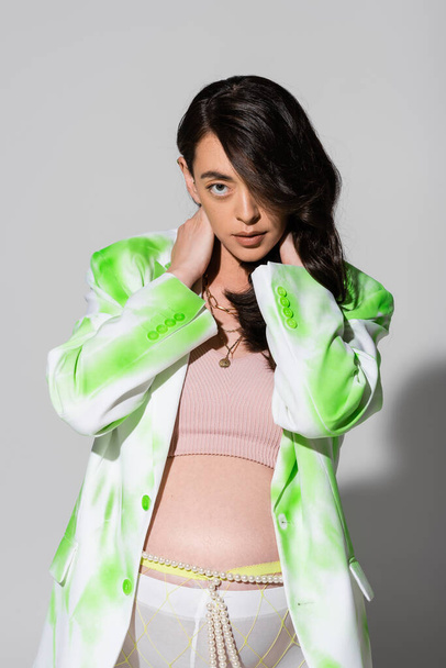 expecting mother with wavy brunette hair posing with hands on neck in green in white blazer, crop top, beads belt and leggings on grey background, maternity fashion concept, expectation - Foto, immagini