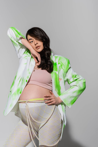 pregnant woman with wavy brunette hair, wearing green and white jacket, crop top, leggings and beads belt standing with hand on hip and looking away on grey background, trendy pregnancy concept - Фото, изображение