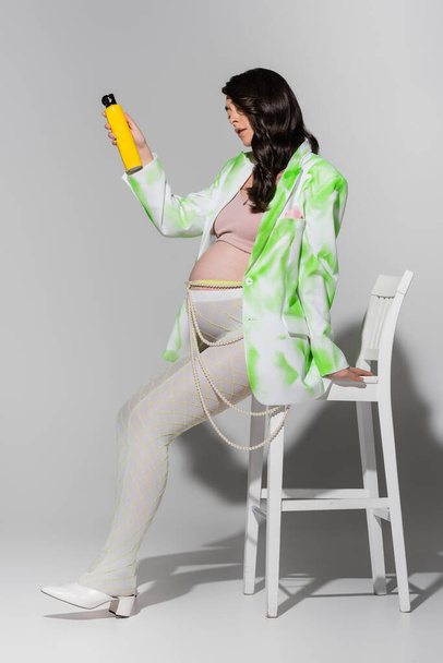 πλήρες μήκος της εγκύου γυναίκας με λαμπερά καστανά μαλλιά, φορώντας κομψό σακάκι, κολάν και χάντρες ζώνη, κάθεται στην καρέκλα και κρατώντας σπρέι μαλλιών σε γκρι φόντο, έννοια της μόδας μητρότητας - Φωτογραφία, εικόνα