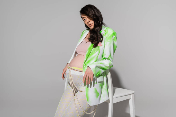 brunette mother-to-be in green and white blazer, leggings, crop top and beads belt looking at camera near chair on grey background, maternity fashion concept, expectation - Foto, imagen