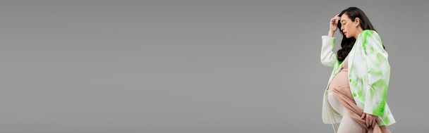 vista lateral da mulher grávida com cabelo morena ondulado posando em blazer verde e branco, top de cultura e leggings com pano de chiffon e cinto de contas isolado em fundo cinza, banner - Foto, Imagem