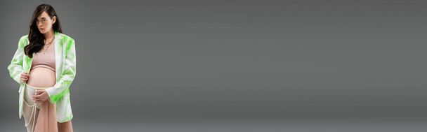 brünette schwangere Frau im trendigen Blazer, bauchfreiem Oberteil, Perlengürtel und Leggings mit beigem Chiffontuch, Blick in die Kamera isoliert auf grauem Hintergrund, Umstandsmodekonzept, Banner - Foto, Bild