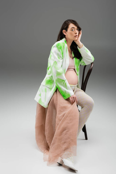 full length of appealing pregnant woman in stylish jacket, crop top and leggings with beads belt and beige chiffon sitting on chair and looking at camera on grey background, maternity fashion concept - Фото, зображення