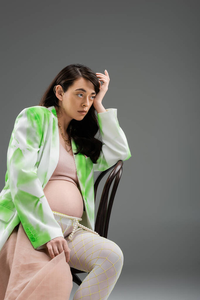 charming brunette mother-to-be in fashionable blazer, crop top and leggings with beads belt and chiffon cloth sitting on chair and looking away on grey background, maternity style concept - Fotó, kép