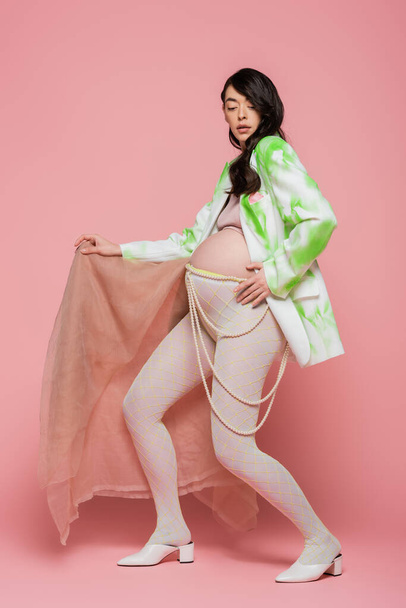 full length of brunette pregnant woman in trendy blazer, crop top, beads belt and leggings posing with beige chiffon cloth on pink background, maternity fashion concept - Фото, зображення