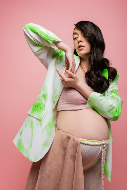 fashionable future mother with wavy brunette hair posing in green and white blazer, crop top and beads belt with chiffon cloth isolated on pink background, pregnancy style concept - Photo, Image