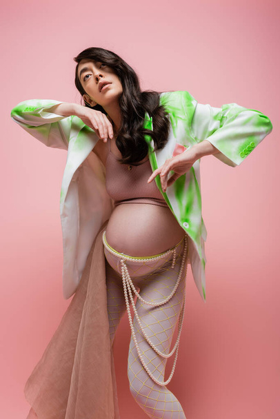 charming mom-to-be posing in green and white jacket, crop top, leggings with beads belt and beige chiffon cloth isolated on pink background, trendy maternity concept, pregnant woman  - Photo, Image