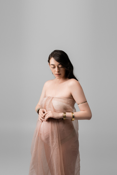 belle-mère gracieuse aux cheveux bruns ondulés, posant en bracelets dorés et drapés en mousseline de soie douce isolés sur fond gris, concept de mode maternité, femme enceinte  - Photo, image