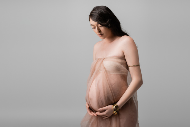 élégante future mère aux cheveux bruns ondulés, debout en chiffon beige et bracelets dorés isolés sur fond gris, concept de mode maternité, femme enceinte  - Photo, image