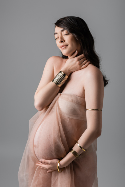 charming and romantic mom-to-be in gentle chiffon draping and golden bracelets standing with closed eyes isolated on grey background, maternity fashion concept, sensuality - Photo, Image