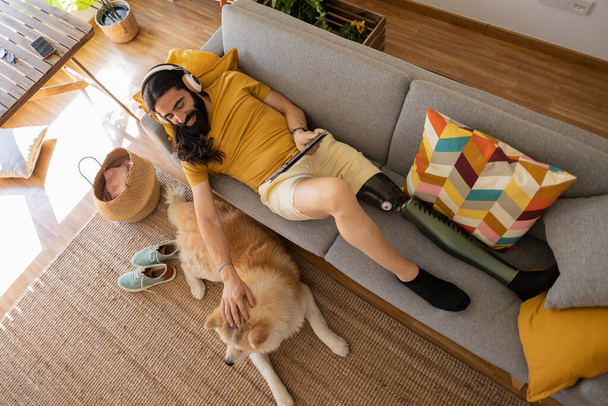 Kanepede müzik dinleyen ve tablet kullanan genç bir adam. Evde köpeğinin yanında. Yapay bir bacağı var., - Fotoğraf, Görsel