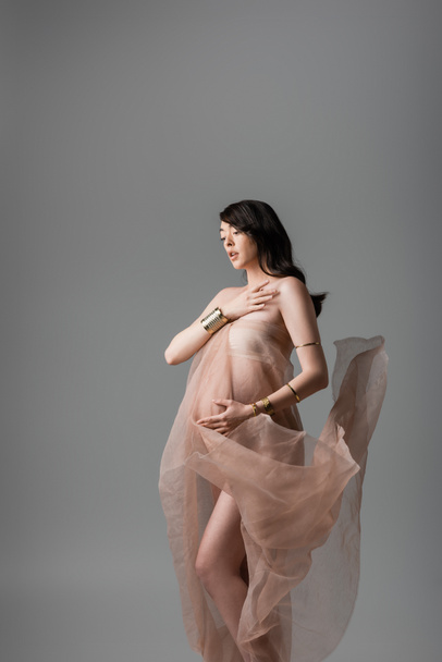 romantyczna matka-to-be dotyka klatki piersiowej podczas pozowania w złote bransoletki i delikatny szyfon zasłony izolowane na szarym tle, koncepcja mody macierzyńskiej, kobieta w ciąży z brzuchem  - Zdjęcie, obraz