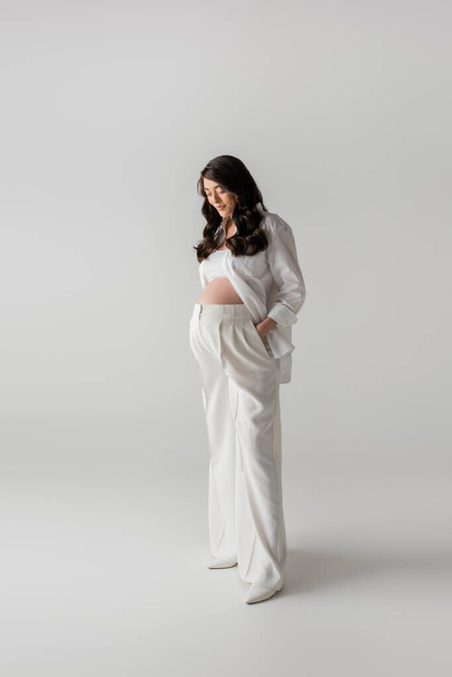 full length of brunette pregnant woman in white shirt and crop top standing with hand in pocket of pants on grey background, fashionable pregnancy concept, expectation, future mother with belly - Photo, Image