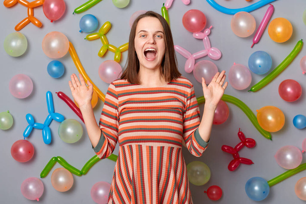 Happy smiling excited woman wearing striped dress standing against gray wall with balloons decoration raised arms screaming with amazement celebrating, festive mood. - Photo, image
