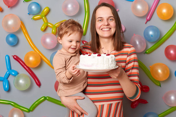 Smiling woman wearing striped dress standing against gray wall with balloons decoration holding her infant baby daughter and birthday cake, delicious dessert. - Photo, Image