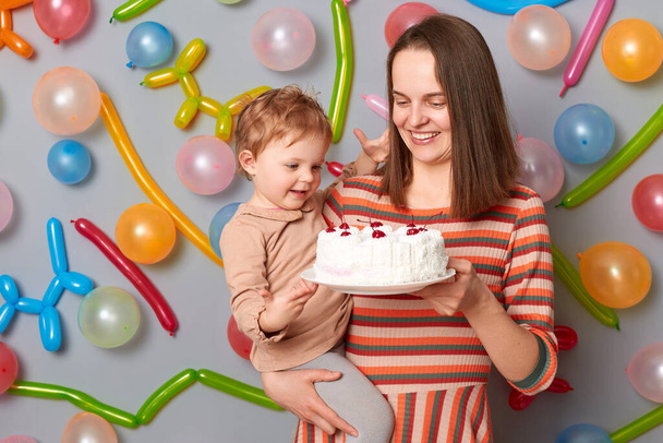 Pleased satisfied woman wearing striped dress standing against gray wall with balloons decoration celebrating birthday with her toddler daughter expressing happiness. - Foto, Imagen