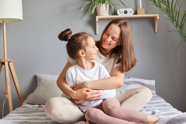Smiling adult woman embraces her cute little girl in their beautiful bedroom, enjoying moment of togetherness and love in their leisure time, hugging expressing love. - Photo, Image